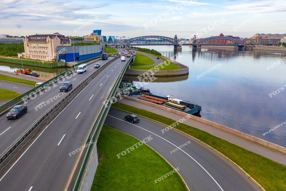 На фотографии изображен Большеохтинский мост в Санкт-Петербурге, один из красивейших мостов города. На переднем плане проходит современная автомобильная магистраль, а вдоль набережной видны прогулочные зоны и пришвартованные суда. Мост соединяет два берега Невы, создавая важную транспортную артерию. Отражения в воде и зелень добавляют гармонии общей композиции.